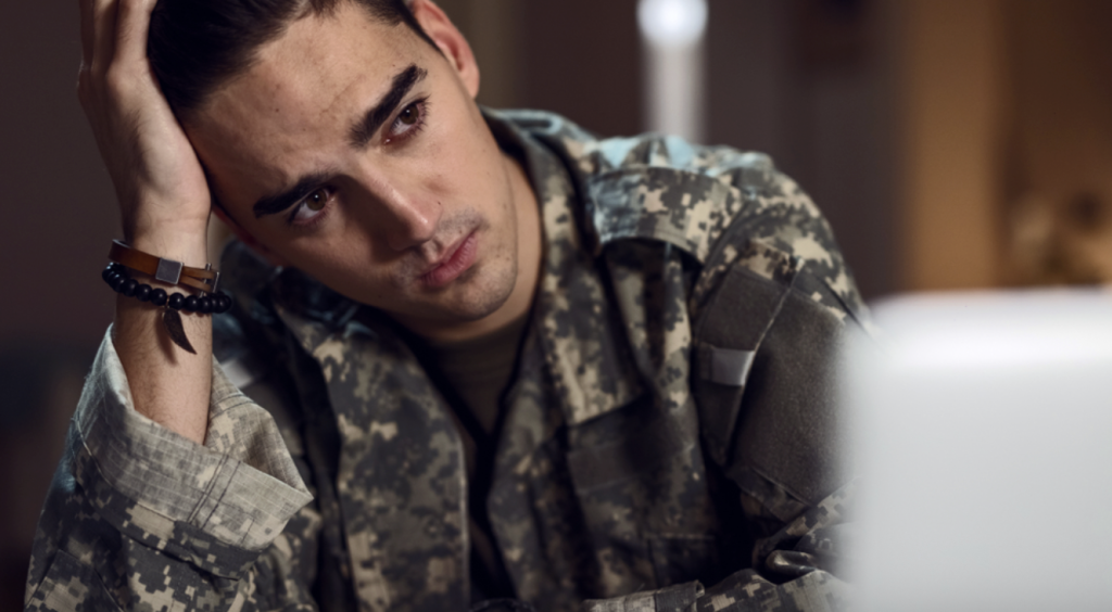 A man in camouflouge sitting at a desk, looking concerned.