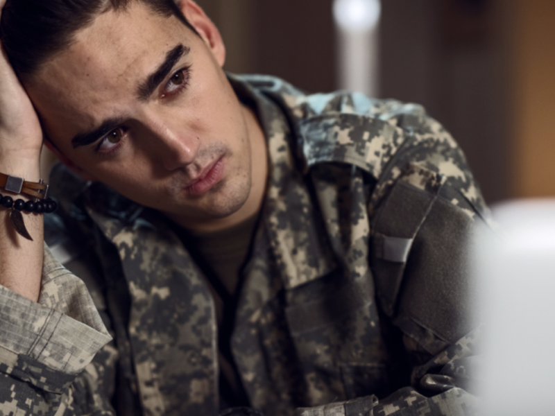 A man in camouflouge sitting at a desk, looking concerned.