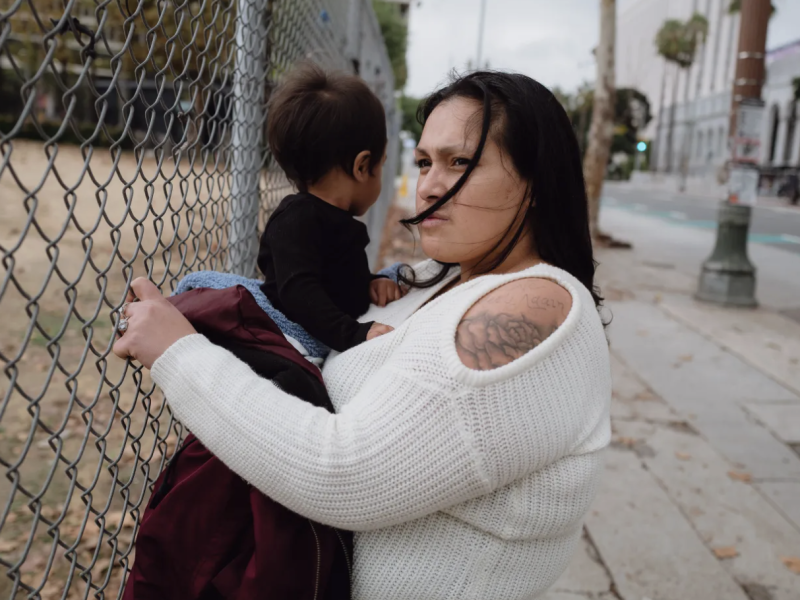 Jazmine Mapes holds her son by the fence where she once lived in a tent while unhoused, just across the street from Los Angeles City Hall. Credit: Zaydee Sanchez for Tradeoffs and The Marshall Project
