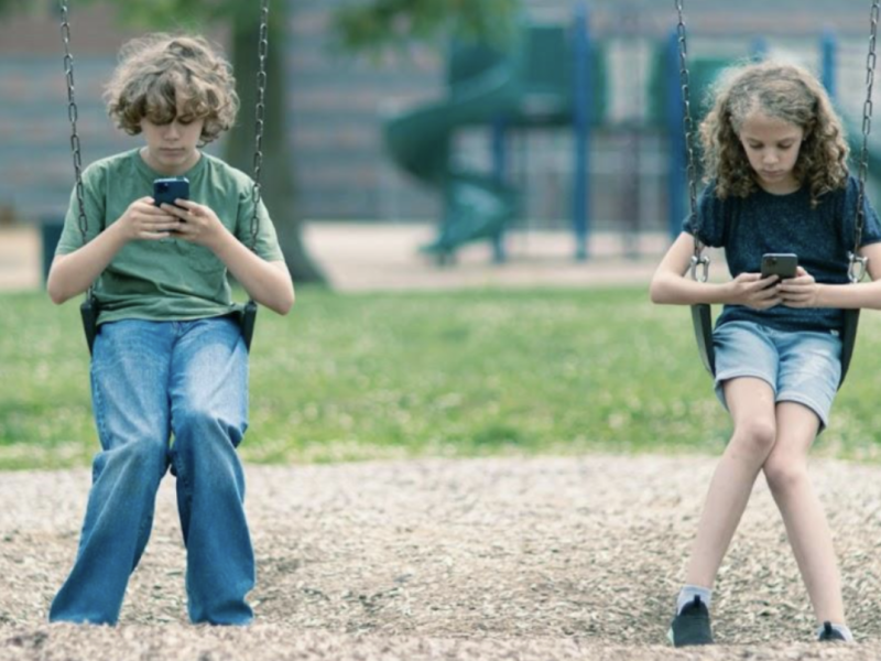 Two children on swings, looking at their phones.