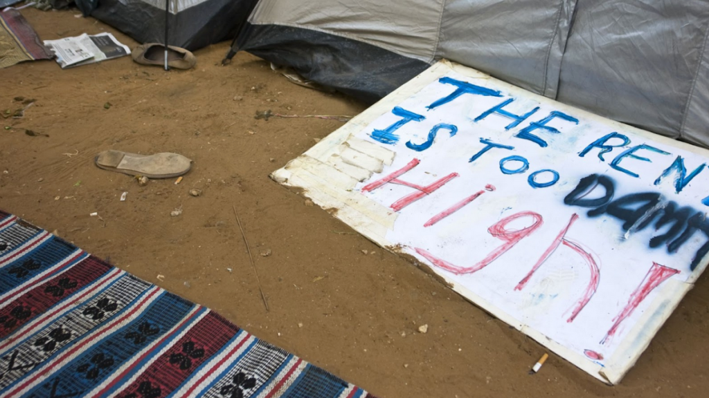 In a homeless encampment, a sign reading "The rent is too damn high!" sits in the dirt, surrounded by tents, blankets and trash.
