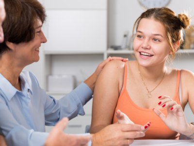 A teenage girl sits at a table, smiling and talking with her parents.