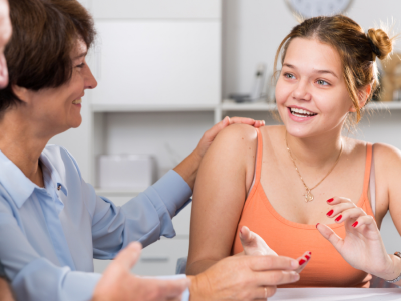 A teenage girl sits at a table, smiling and talking with her parents.