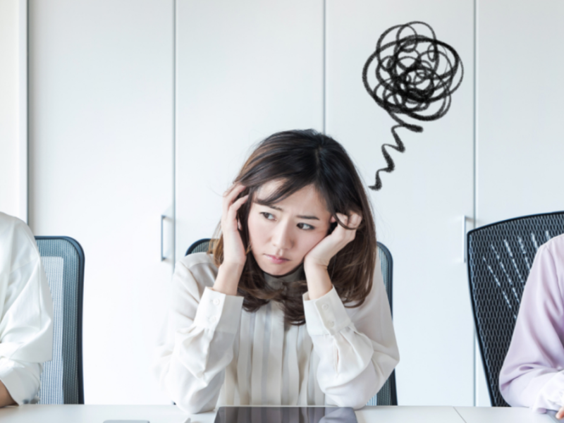 A woman in professional clothes sits at a table, frowning and staring off into space as other women around her work on their computers.