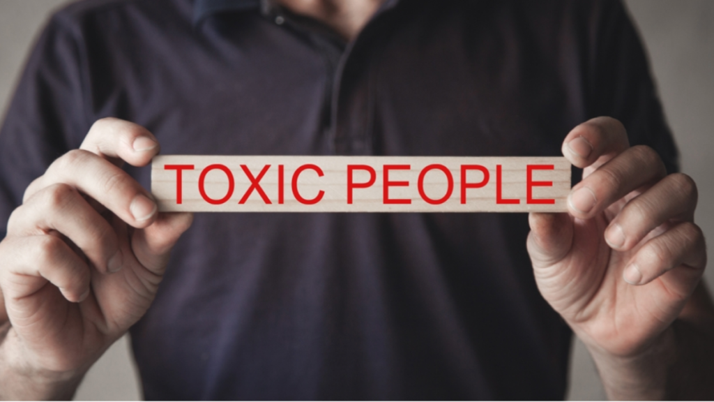 A man holds a wooden block with "toxic people" printed on it in red, capitalized letters.