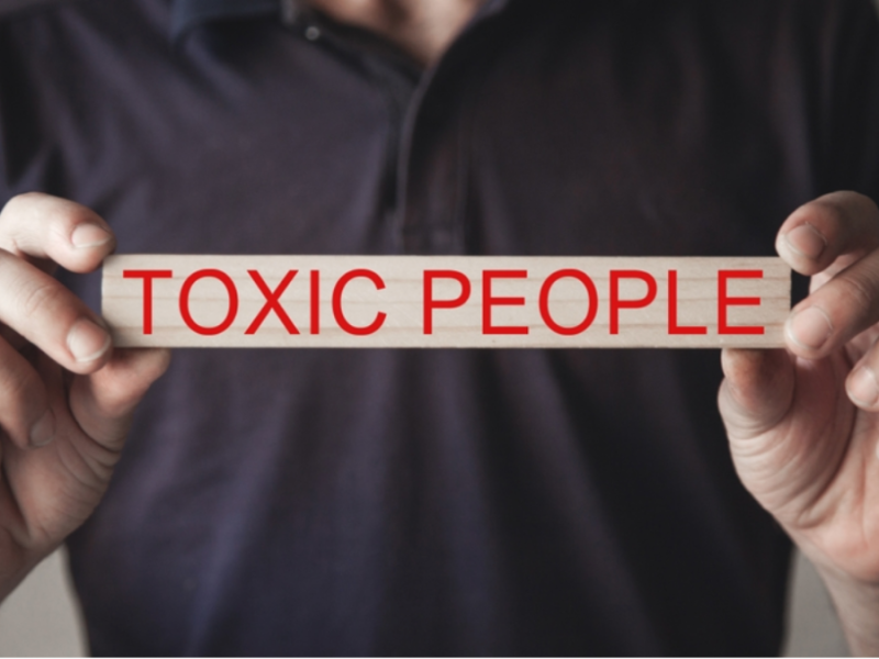 A man holds a wooden block with "toxic people" printed on it in red, capitalized letters.