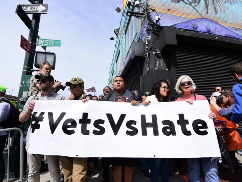 A group of protesters hold a sign reading "#VetsVsHate"