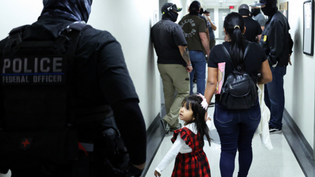 A little girl is led down a hallway by her mother. The two are surrounded by multiple masked federal agents.