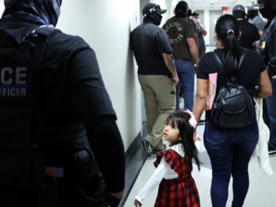 A little girl is led down a hallway by her mother. The two are surrounded by multiple masked federal agents.