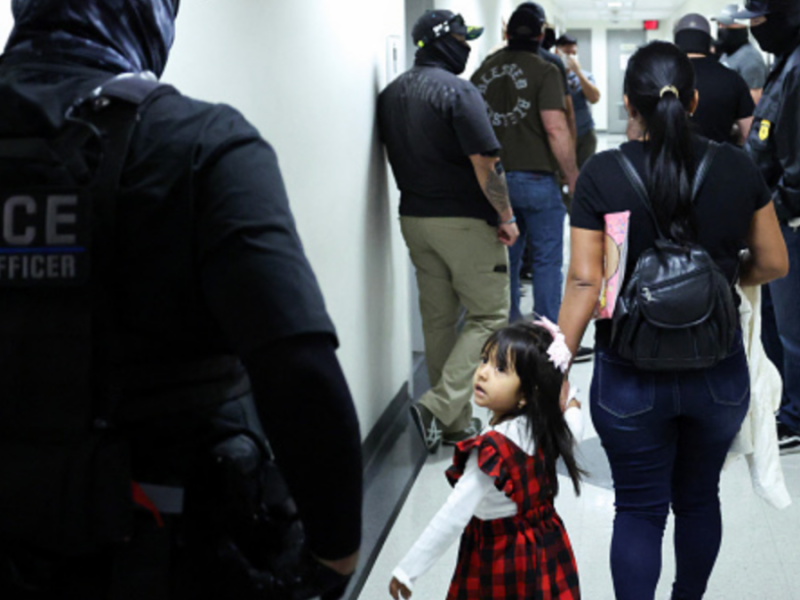 A little girl is led down a hallway by her mother. The two are surrounded by multiple masked federal agents.