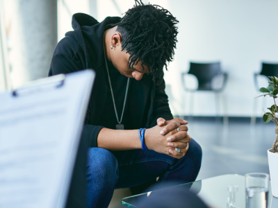 A teen boy sits in a therapist's office with his head down, looking at the ground.