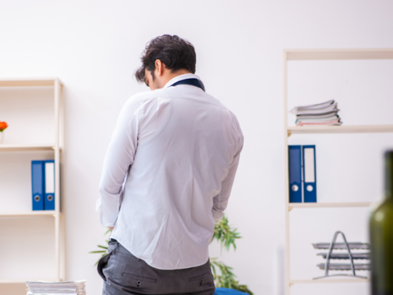 A man stands in an office, with his back to the camera. An empty wine bottle sits on a table behind him