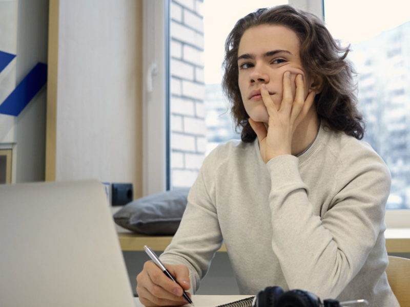A young man sits at a table with a laptop, notebook and pen, resting his head on his hand