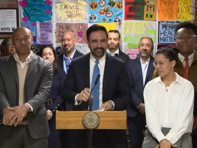 New York City Major Zohran Mamdani stands at a podium, surrounded by a group of people
