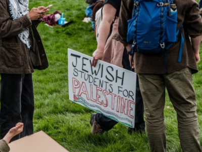 A group of protesters stands in a park, one holding a sign reading "Jewish Students for Palestine."