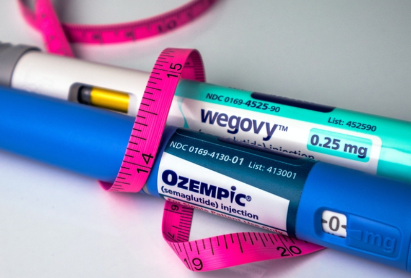 An Ozempic injection pen and Wegovy injection pen sit on a table, tied up by a measuring tape