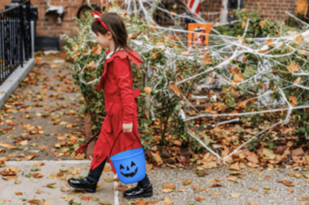 A young girl wearing a devil costume for halloween walks down the sidewalk, holding a blue pumpkin bucket.