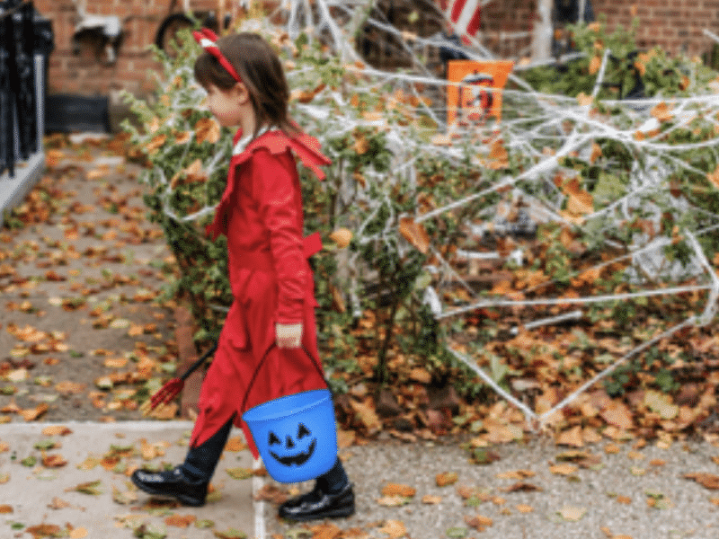 A young girl wearing a devil costume for halloween walks down the sidewalk, holding a blue pumpkin bucket.